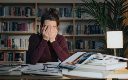 Eine Frau sitzt an ihrem Schreibtisch, auf dem viel Arbeit liegt, und hält sich die Augen zu.