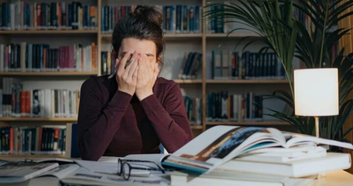 Eine Frau sitzt an ihrem Schreibtisch, auf dem viel Arbeit liegt, und hält sich die Augen zu.