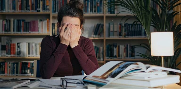 Das Bild zeigt eine Frau, die an einem Schreibtisch sitz und sich die Augen zuhält, weil vor ihr ein Berg an Büchern, Zeitschriften und vieles mehr liegt. 