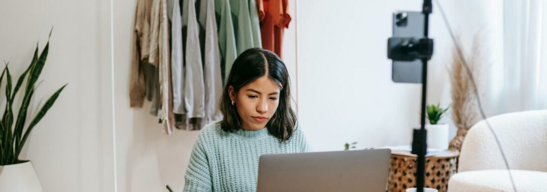 Eine junge Frau sitzt an ihrem Schreibtisch und schreibt auf ihrem Blog und ihrem Laptop.