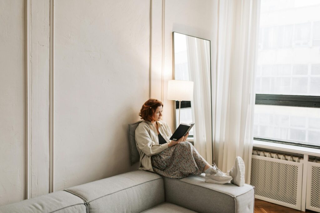 Eine Frau sitzt gemütlich am Rande einer Couch und liest ein Buch. 