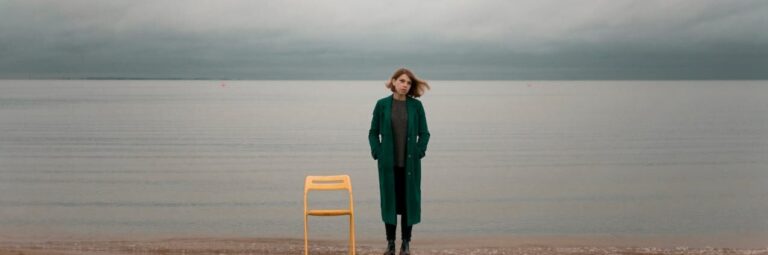 Nachdenkliche Frau neben leerem Stuhl am Strand – Metapher für warum sich mein Leben nicht verändert