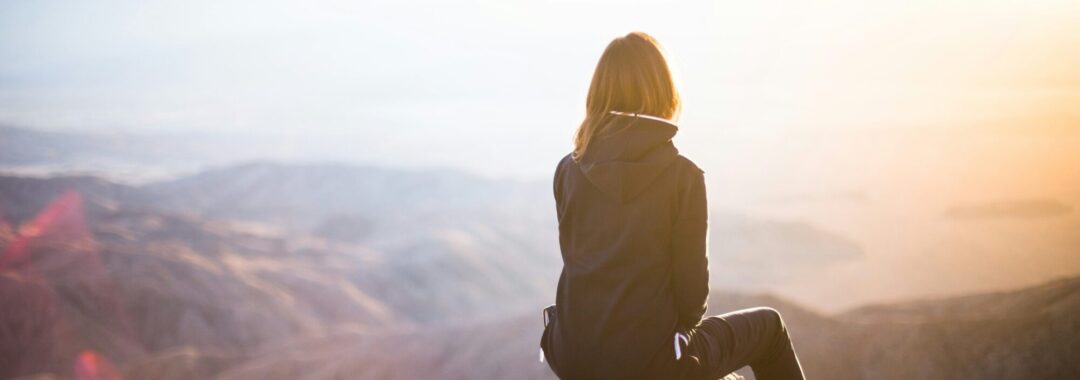 Frau sitzt auf einem Felsen Frau sitzt auf einem Felsen und blickt in die Ferne – symbolisch für das Innehalten und Reflektieren