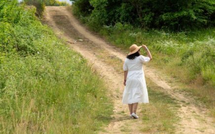 Eine Frau im weißen Kleid und einem eleganten Sonnenhut spaziert auf einem Feldweg, um ihre Gedanken zu ordnen, den Alltag hinter sich zu lassen und den Kopf komplett frei zu bekommen.