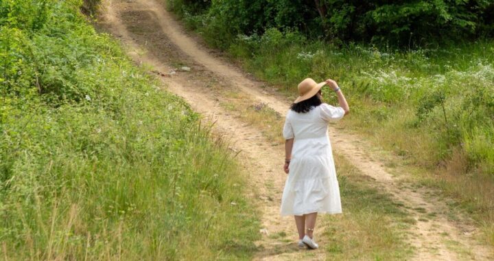 Eine Frau im weißen Kleid und einem eleganten Sonnenhut spaziert auf einem Feldweg, um ihre Gedanken zu ordnen, den Alltag hinter sich zu lassen und den Kopf komplett frei zu bekommen.