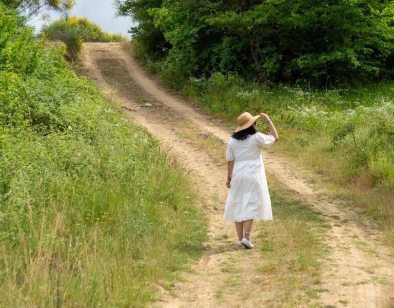 Eine Frau im weißen Kleid und einem eleganten Sonnenhut spaziert auf einem Feldweg, um ihre Gedanken zu ordnen, den Alltag hinter sich zu lassen und den Kopf komplett frei zu bekommen.